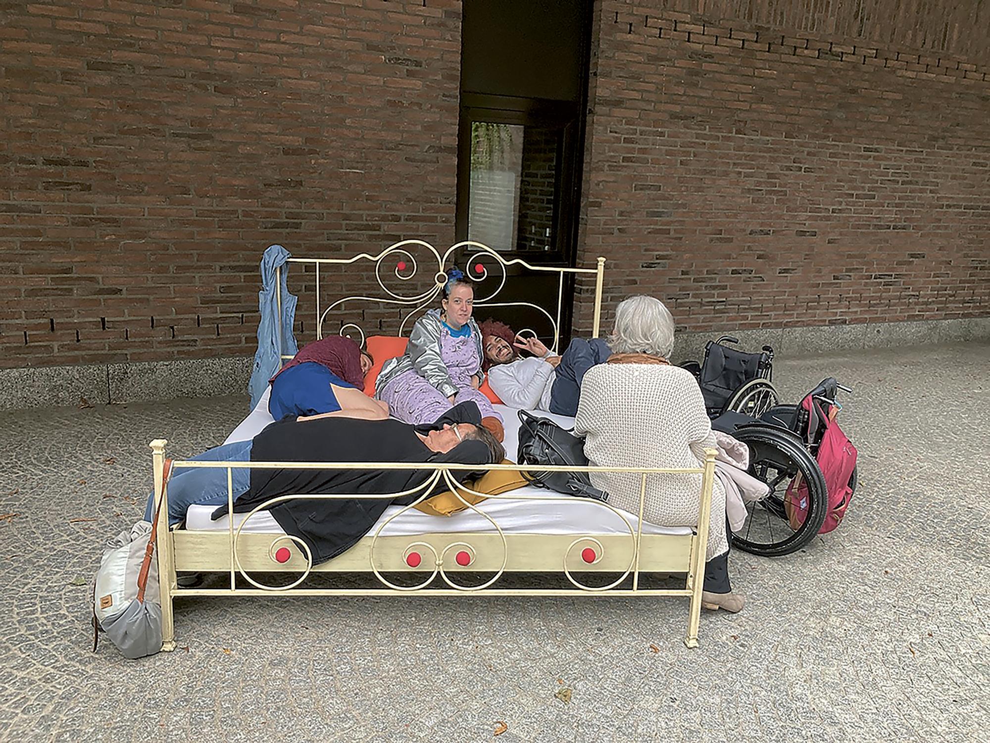 A cobblestoned floor in front of a brick-walled gallery. Angela Alves’ double bed with a pale beige frame and red embellishments stands in front of the wall. On the bed are a mattress covered with a white sheet and pillows in orange and yellow. Five people are lying or sitting on the bed; among them are Zurich artists Nina Muehlemann and Edwin Ramirez. Their wheelchairs are standing next to the bed.