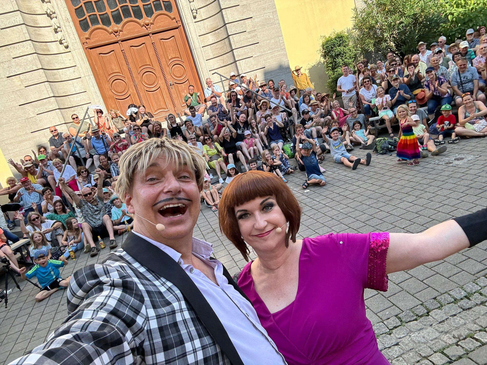 Ein Mann mit blonder Perücke und Schnurrbart sowie eine Frau mit rotbrauner Pagenfrisur im pinken Kleid machen ein Selfie, beide tragen Headsets. Hinter ihnen applaudiert und winkt ein großes Publikum, das im Freien auf Stufen und Pflaster vor einem Portal sitzt.
