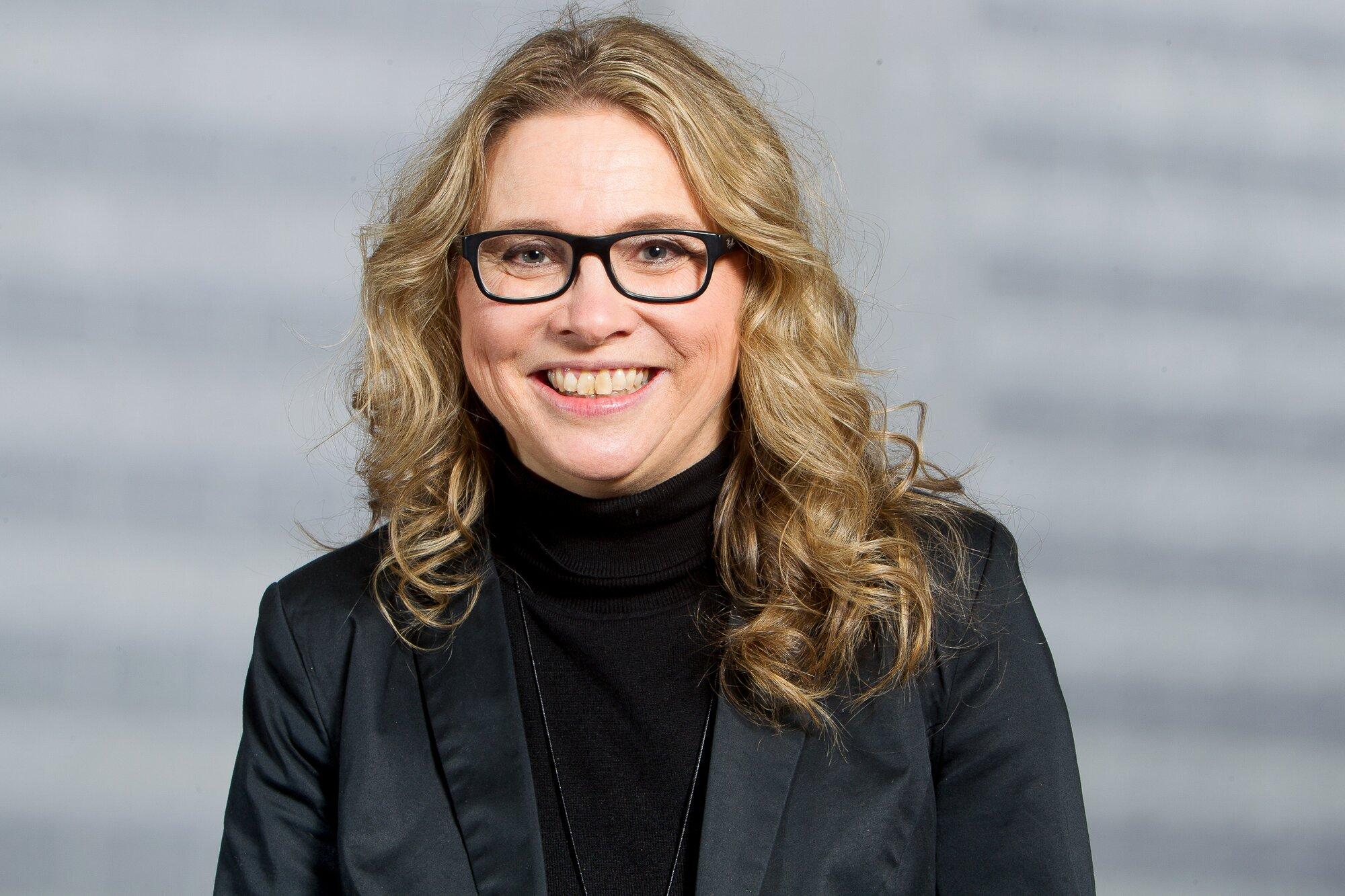 Porträt einer lächelnden Frau mit langen, welligen blonden Haaren und schwarzer Brille, die einen schwarzen Rollkragenpullover und einen schwarzen Blazer trägt, vor einem unscharfen grauen Hintergrund.