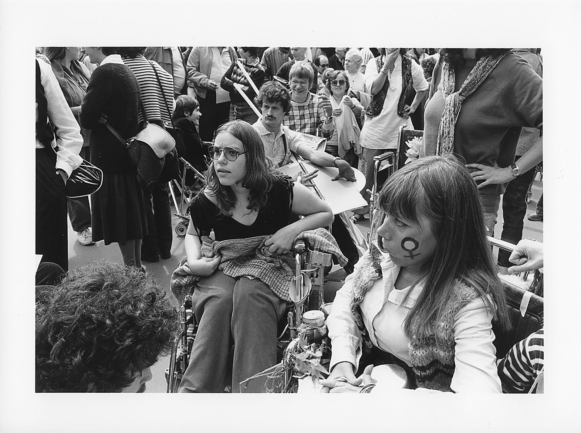 Auf dem Foto sieht man eine Menschenansammlung an der großen schweizweiten Behinderten-Demonstration in Bern am 20. Juni 1981. Ein Mann im Hintergrund hält ein Transparent auf dem Schoß. Im Vordergrund des Fotos ist Ursula Eggli. Sie benutzt einen Rollstuhl und hat ein feministisches Symbol auf die Wange gemalt. Neben ihr sitzt die Behindertenaktivistin und Künstlerin Cornelia Nater. Das Foto wurde von der Fotografin und Aktivistin Helga Leibundgut gemacht. Sie war eine Bekannte von Ursula Eggli und Mitglied der Frauenbefreiungsbewegung FBB. (Schweizerisches Sozialarchiv, F 5110-Fc-102)