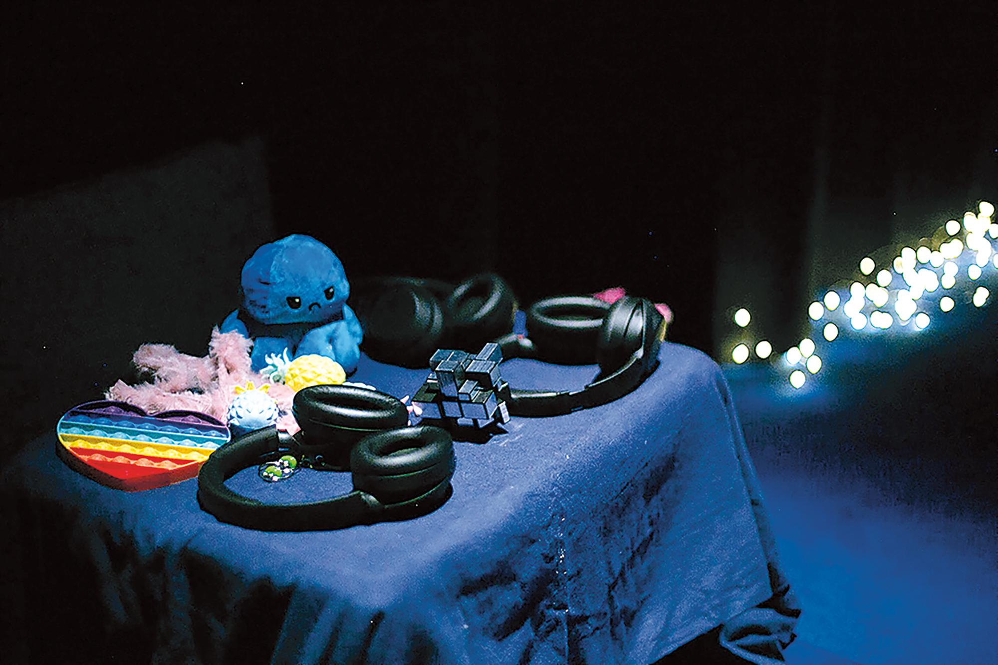 A dimly lit table covered with blue fabric. On it are placed stim toys, plushies, and noise-cancelling headphones. In the background a string of fairy lights is visible. It is an image of the relaxed space, part of the piece “Creature Comforts” by Criptonite and Symbionts (Tanzhaus Zurich, 2023).
