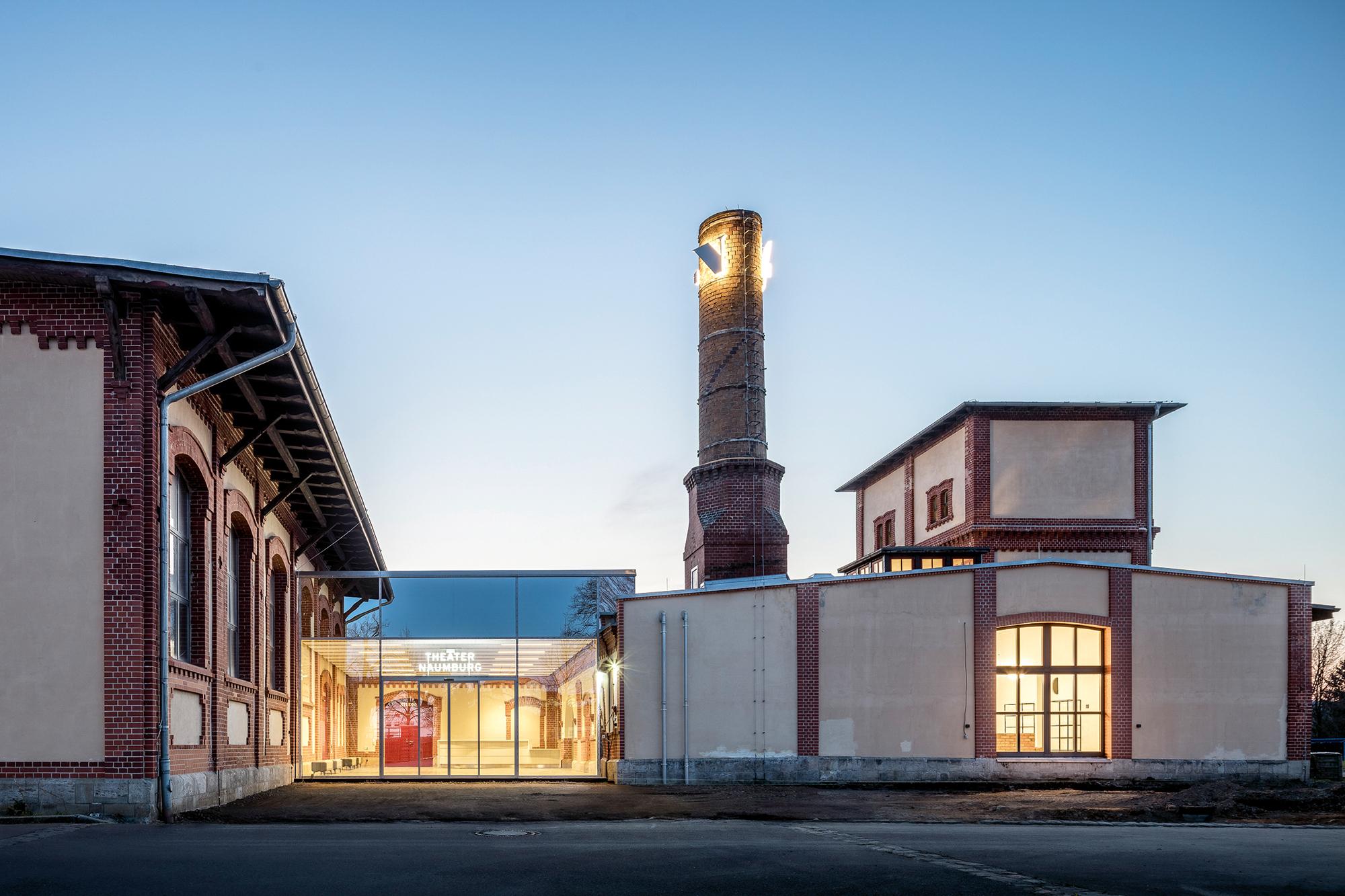 So geht man fortan auf das Theater Naumburg zu. Die Glasfassade zwischen den Gebäudeteilen ist nach außen hin verspiegelt. Am alten Schornstein prangt das Kürzel TN als Leuchtschrift. Rechts neben dem Schornstein, am Kühlturm, fehlt hier noch das Emaille-Schwein des Künstlers Moritz Götze.