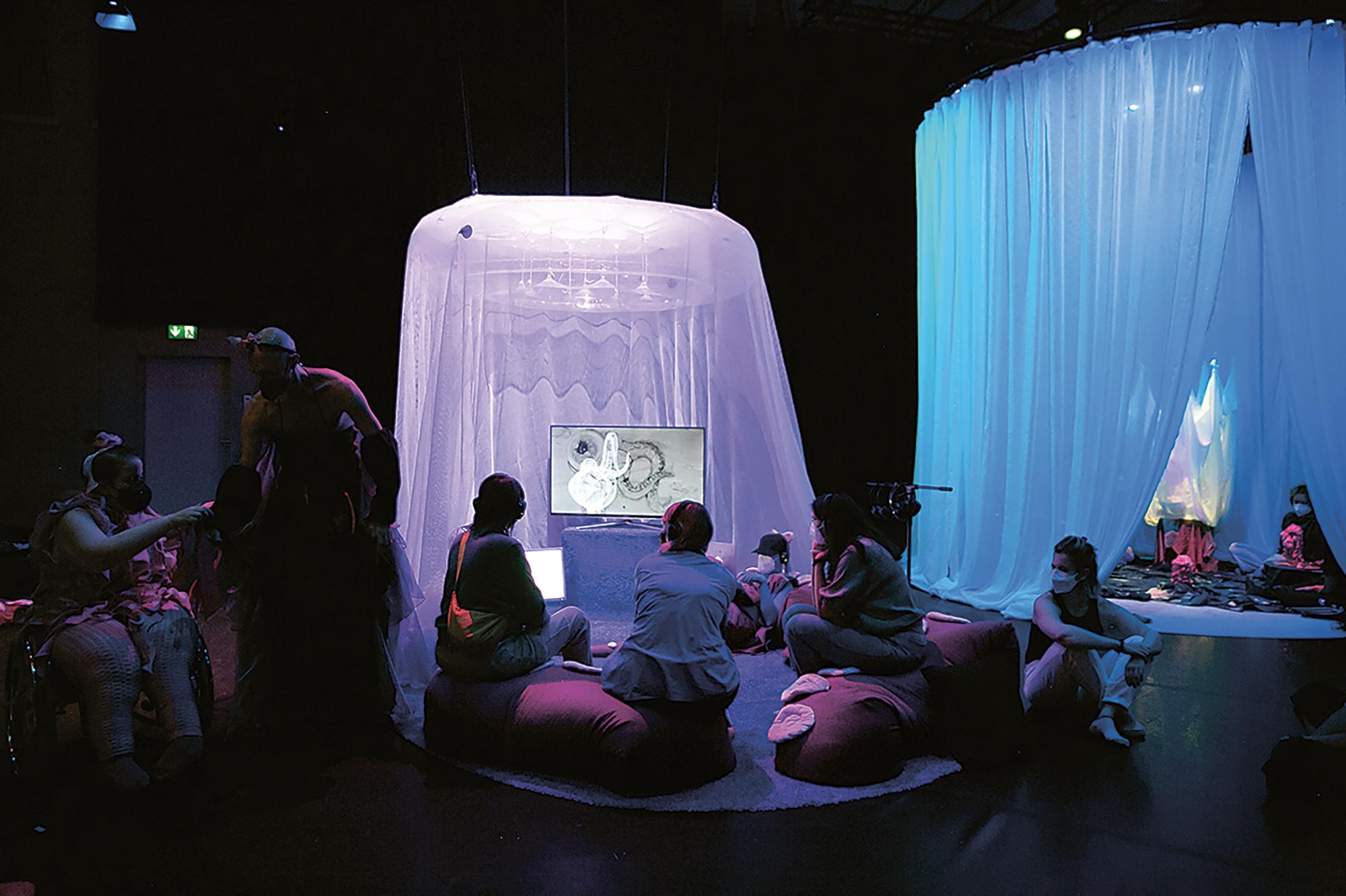 A dimly lit space, in the middle is a purple-lit round carpeted area, halfway enclosed with a mesh curtain. In front of the curtain, a screen displays Kamran Behrouz’ video installation. Kamran is sitting next to it, typing into a computer. Audience members sit on the floor or on magenta tentacle beanbags and watch the video or just chill out. On the right side of the purple-lit area is a similar mostly-curtained-off area lit in blue. On the left side of the purple-lit area are performers Alessandro Schiattarella and Nina Muehlemann, dancing together.