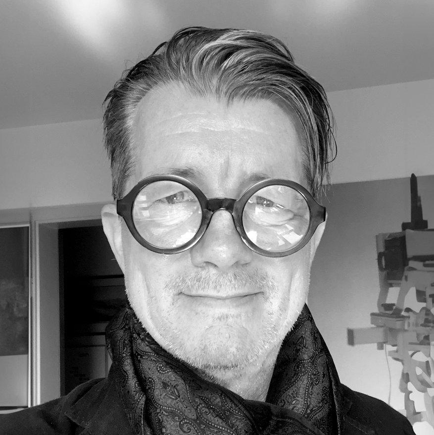 Schwarz-Weiß-Porträt eines Mannes mit runder Brille, dunklem Schal und Bart, im Hintergrund eine holzartige Konstruktion und eine Zimmerecke.