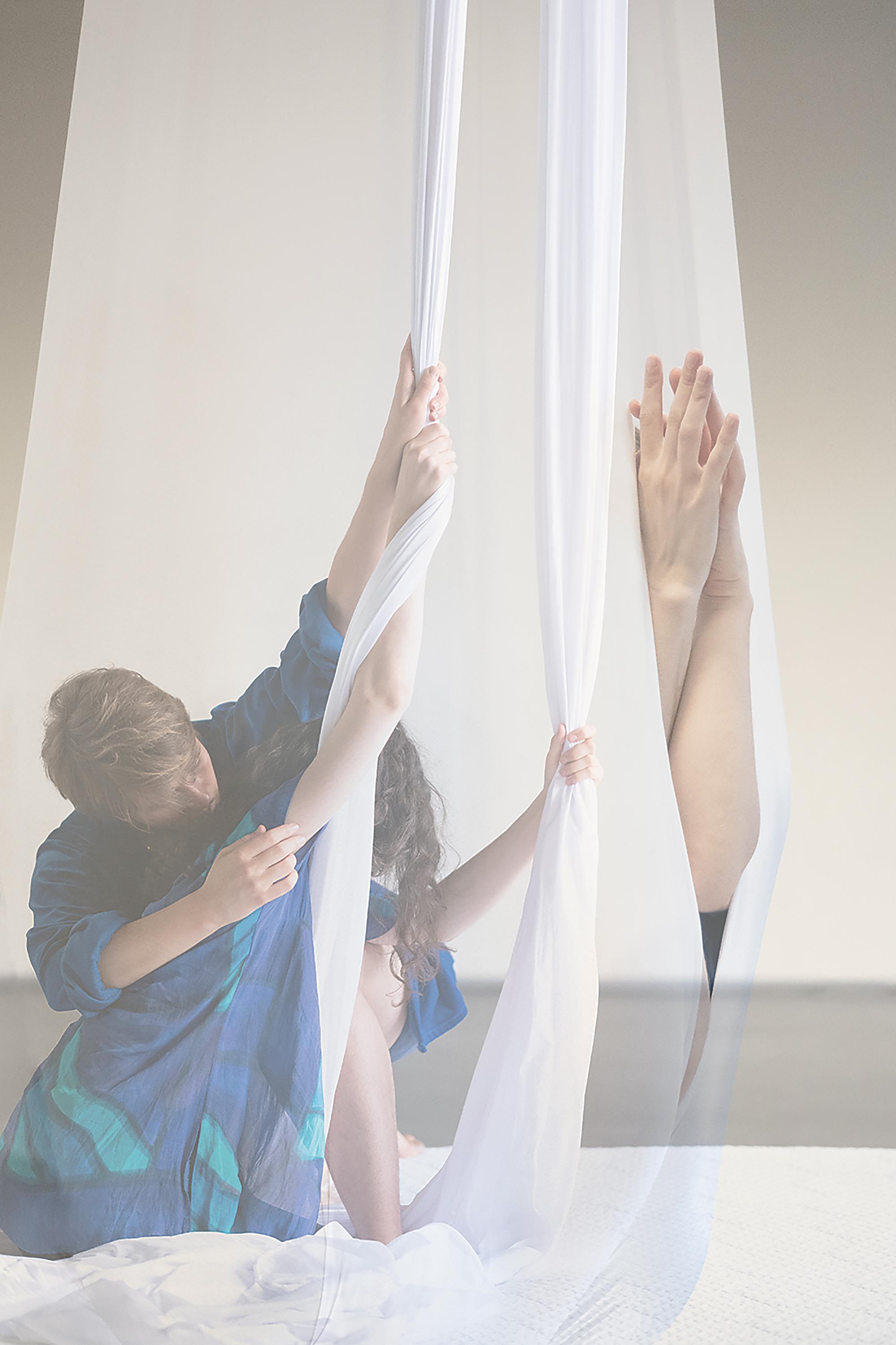 Die Fotografie zeigt Irene Giró und Fia Neises. Die zwei Performer*innen von WITH OR WITHOUT YOU. Beide sind weiß und behindert. Beide werden weiblich und nicht-behindert gelesen. Irene hat braune, dichte, lange Locken und ist feingliedrig. Fia hat dunkelblondes, glattes, ohrlanges Haar. Ihr Körper ist größer und weicher als der von Irene. Die Fotografie zeigt beide im weißen „Silk“. Ein 14 Meter langes Tuch, das in der Mitte an der Decke aufgehängt wird. Die beiden Enden hängen schwer bis zum Boden und die Protagonist*innen wickeln, klettern, schwingen, hängen und halten sich darin. Fia ist blind und hat Irene gefragt, was sie auf dem Bild tun. Dann haben sie sich erinnert: Irene: Wunderschön. Alles wirkt ein wenig wie eine Hochzeitseinladung. Viel weißer fließender Stoff und Sinnlichkeit. Xenia hat zwei Bilder schemenhaft übereinandergelegt.  Fia: Welche? Irene: In dem Moment auf dem hinteren Bild sind wir beide auf der weichen Matte, eng zusammen und beide im Kontakt mit dem Silk.  Fia: Ich erinnere mich. Der Moment war ruhig. aber sehr schön. Ich habe mit meiner Hand versucht nachzuvollziehen, wie deine Hand oben das Silk anfasst. Dein Arm reichte nach oben und dein Gesicht war sehr zuhörend und spürend Richtung Boden geneigt. Du hattest es auf meinem aufgestellten Bein abgelegt.  Irene: Ich sehe deine Hand an der Wurzel von meinem Arm. Außerdem die Krone von deinem Kopf mit den hellblonden Strähnen, die fast meinen Rücken berührt. Mein Kopf verschwindet hinter dem linken Silk. Das andere Silk ist rechts daneben. Es ist genauso gespannt wie das linke Silk, weil es von deiner Hand nach unten gezogen wird. Darüber liegt das Bild von uns beiden im Kokon.  Fia: Wenn wir uns ganz im Silk verstecken. Beide Körper in einer Kugel. Gehalten von einem stabilen Knoten unter unserem Kokon.  Irene: Das Bild zeigt nur unsere Hände, die sich in einer Berührung vorsichtig aus einem Spalt im Kokon wagen. Der Kokon mit unseren Händen liegt blass, wie eine Erinnerung, über unseren beiden rastenden und tastenden Körpern.