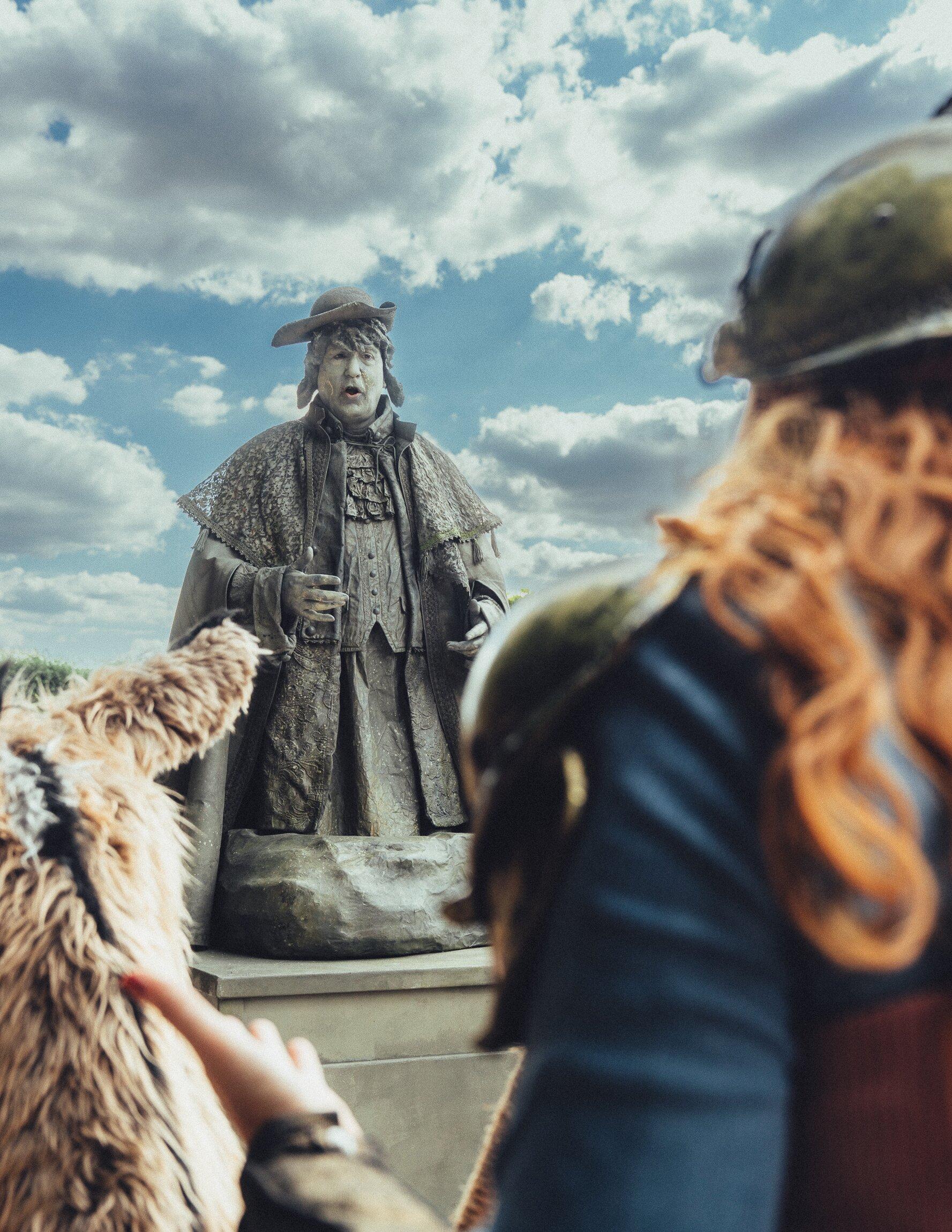 Ein Darsteller mit grau bemaltem Gesicht und steinfarbenem Kostüm aus Dreispitz, Umhang und Rüschenweste steht wie eine Statue auf einem Sockel und singt oder spricht. Im Vordergrund, unscharf, eine Person mit Helm und langen roten Haaren sowie ein zottiges Fellwesen.