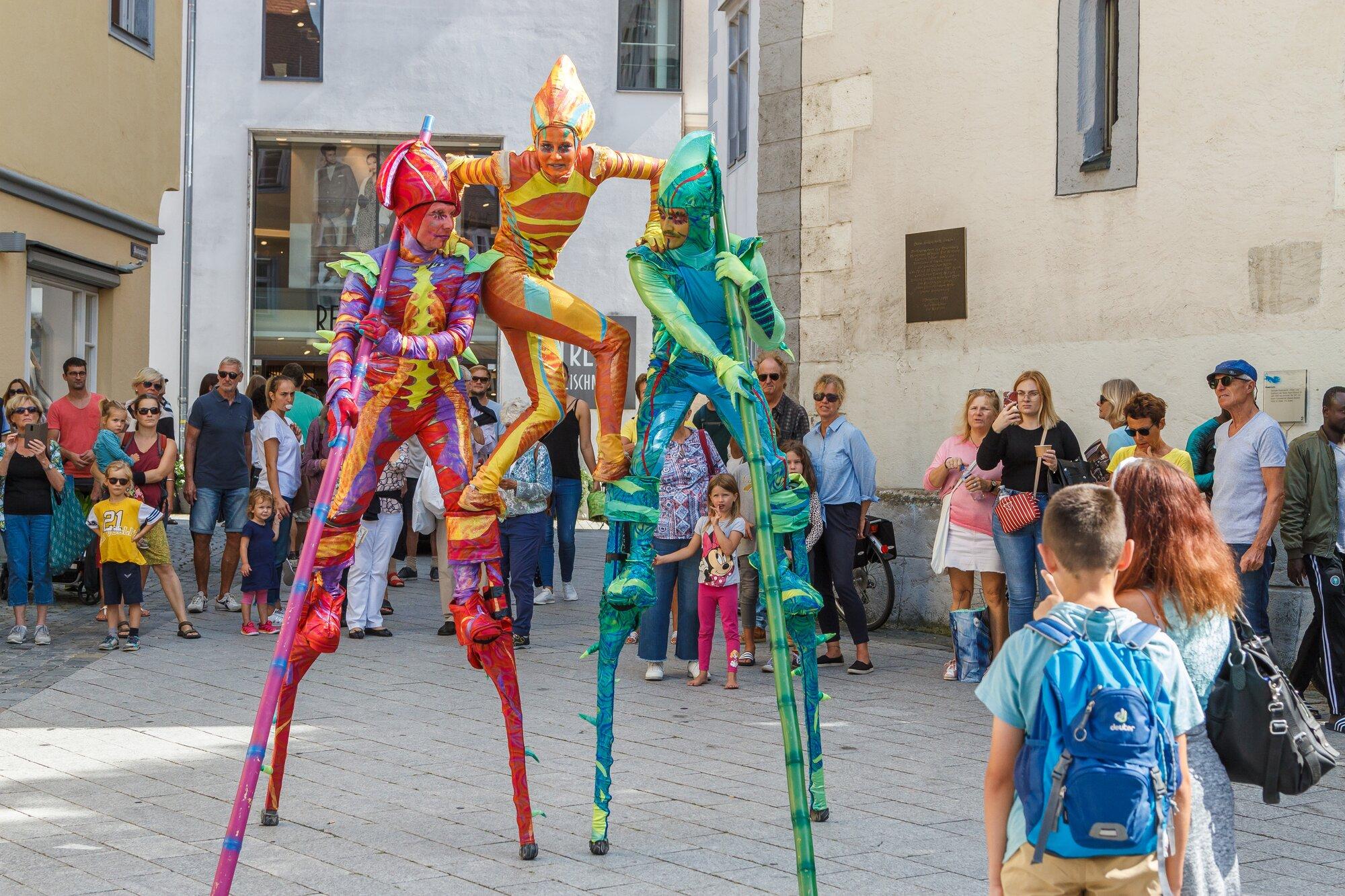 Drei Stelzenläufer in knallbunten Ganzkörperkostümen in Rot, Orange und Grün mit spitzen Kopfbedeckungen stehen auf einem Platz vor Altstadtfassaden; die mittlere Figur stützt sich auf die beiden anderen. Zahlreiche Erwachsene und Kinder schauen zu.