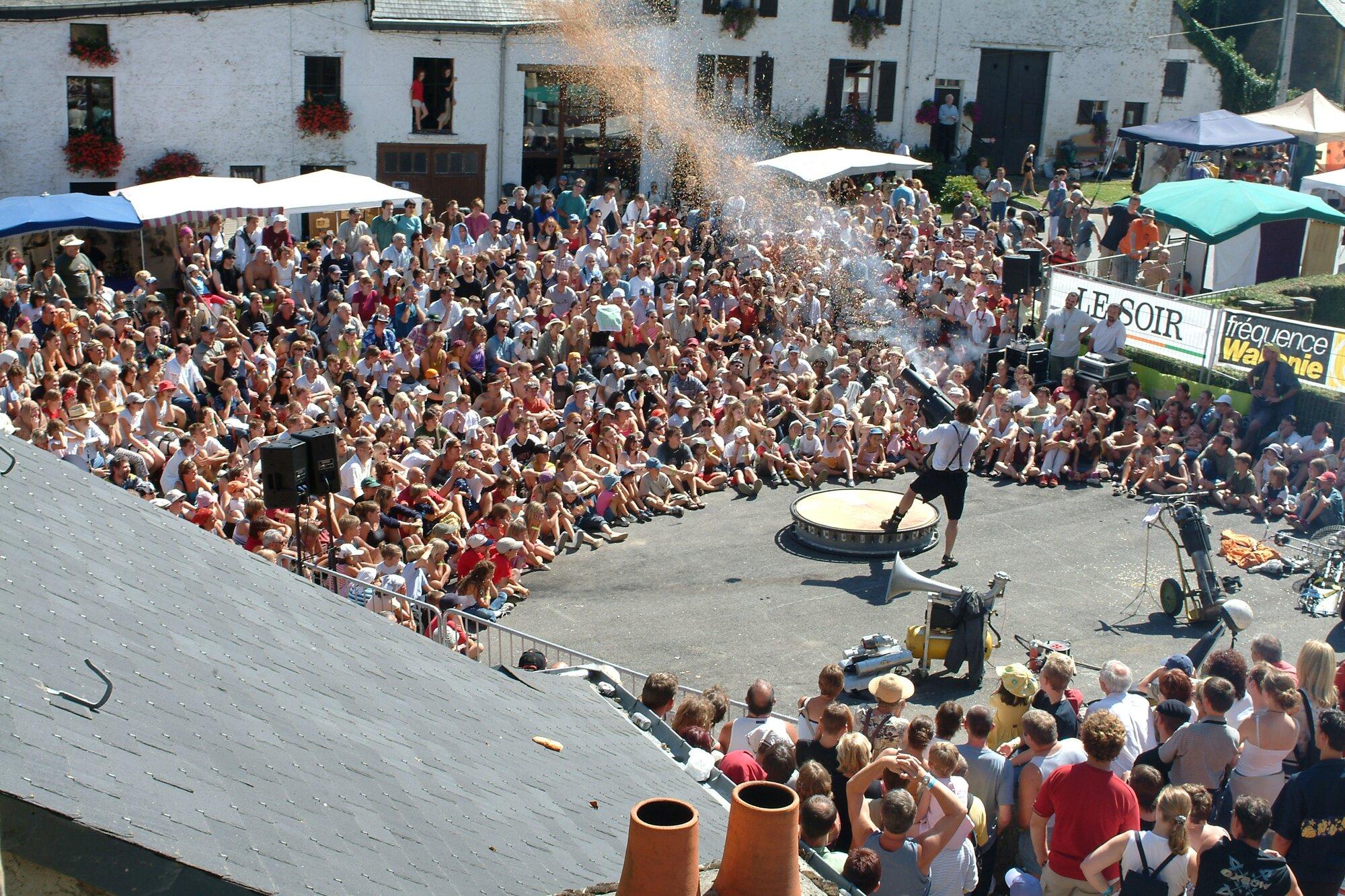 Von einem Dach aus gesehen sitzt und steht eine dichte Menschenmenge im Halbkreis um einen Platz, auf dem ein Mann in kurzer Hose und Hosenträgern eine Kanone auf die Zuschauer richtet. Aus ihr steigt eine Wolke feiner Partikel auf; daneben stehen eine runde Scheibe und technisches Gerät.
