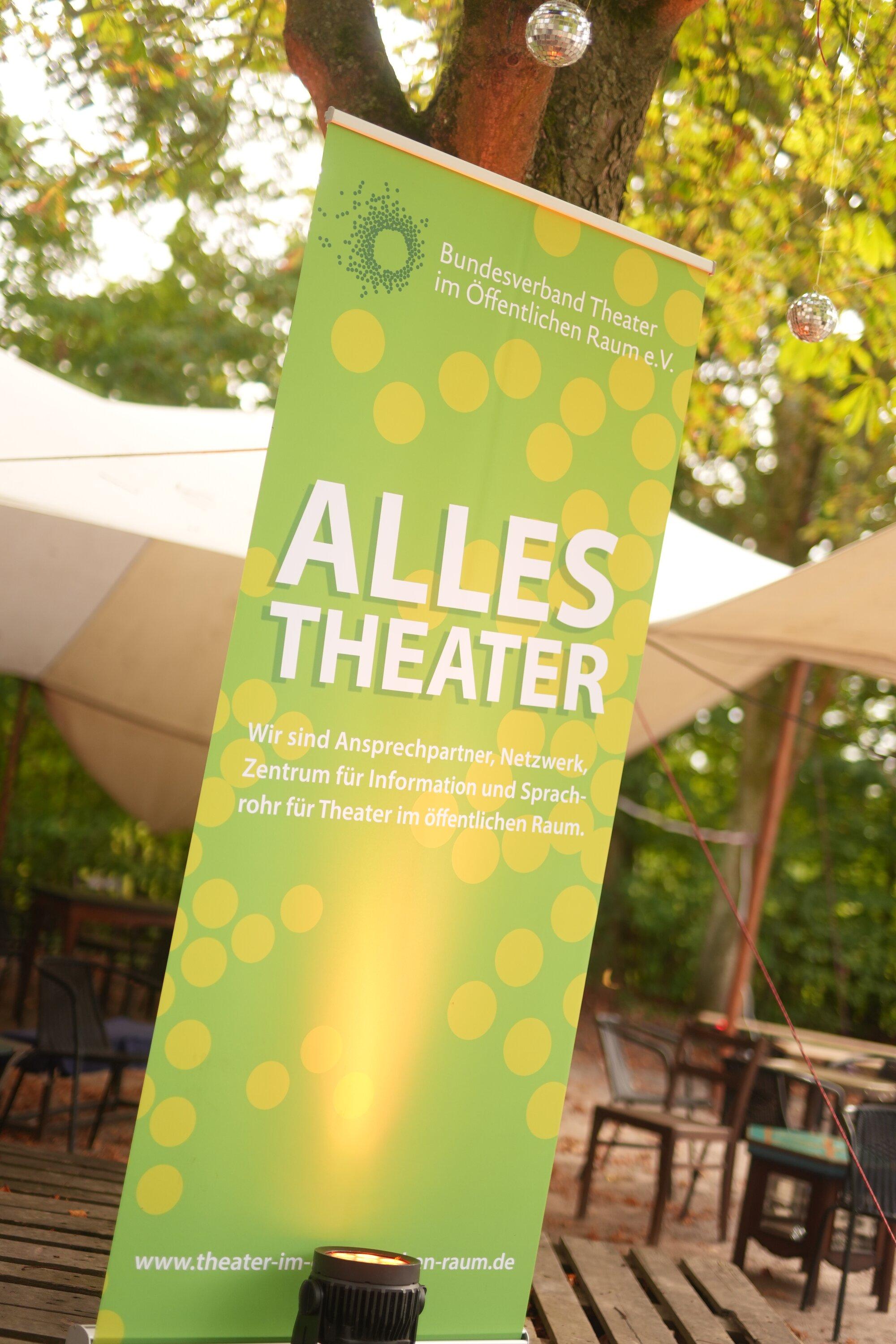 Ein grünes Roll-up-Banner mit gelben Punkten und der Aufschrift „Alles Theater" des Bundesverbands Theater im Öffentlichen Raum steht im Freien, von unten angestrahlt. Dahinter sind ein helles Zeltdach, Bäume mit Spiegelkugeln und Stühle auf Sandboden zu sehen.