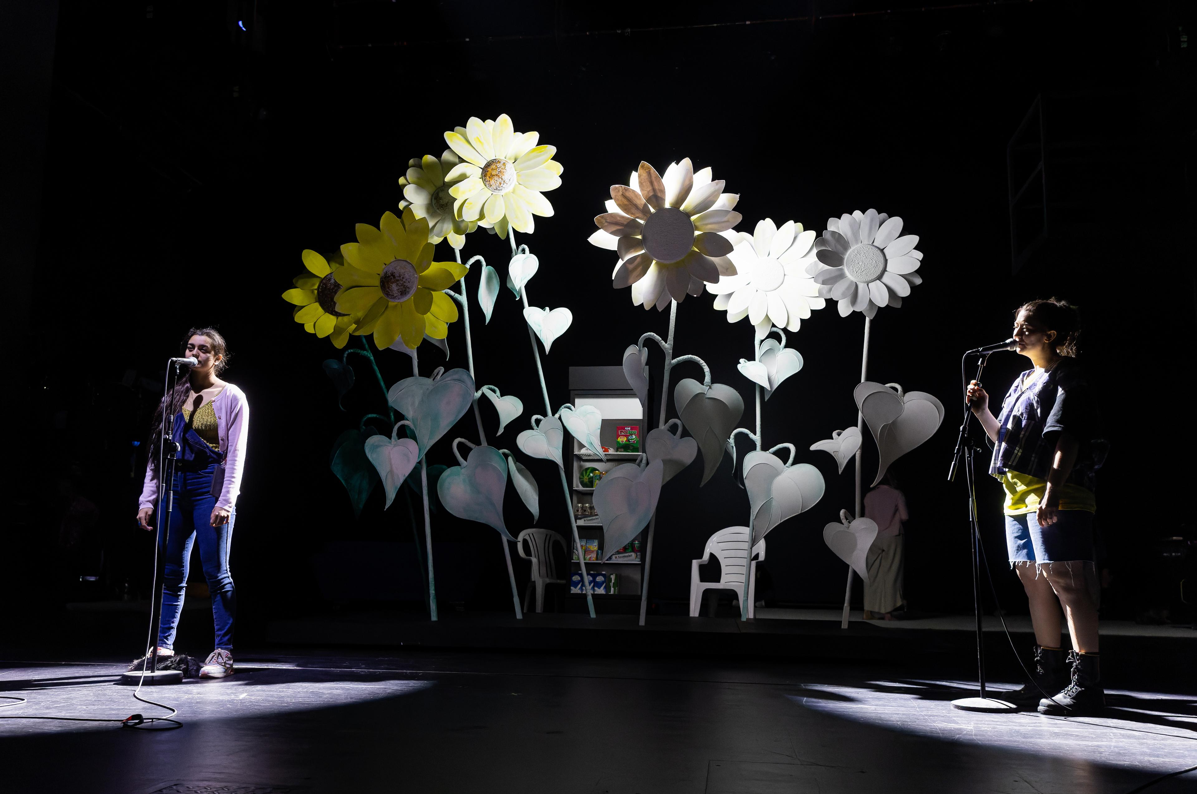 Melanie Sidhu als Gwen und Nina Carolin als Charles vor Sonnenblumen, die so groß sind, wie Bäume.
