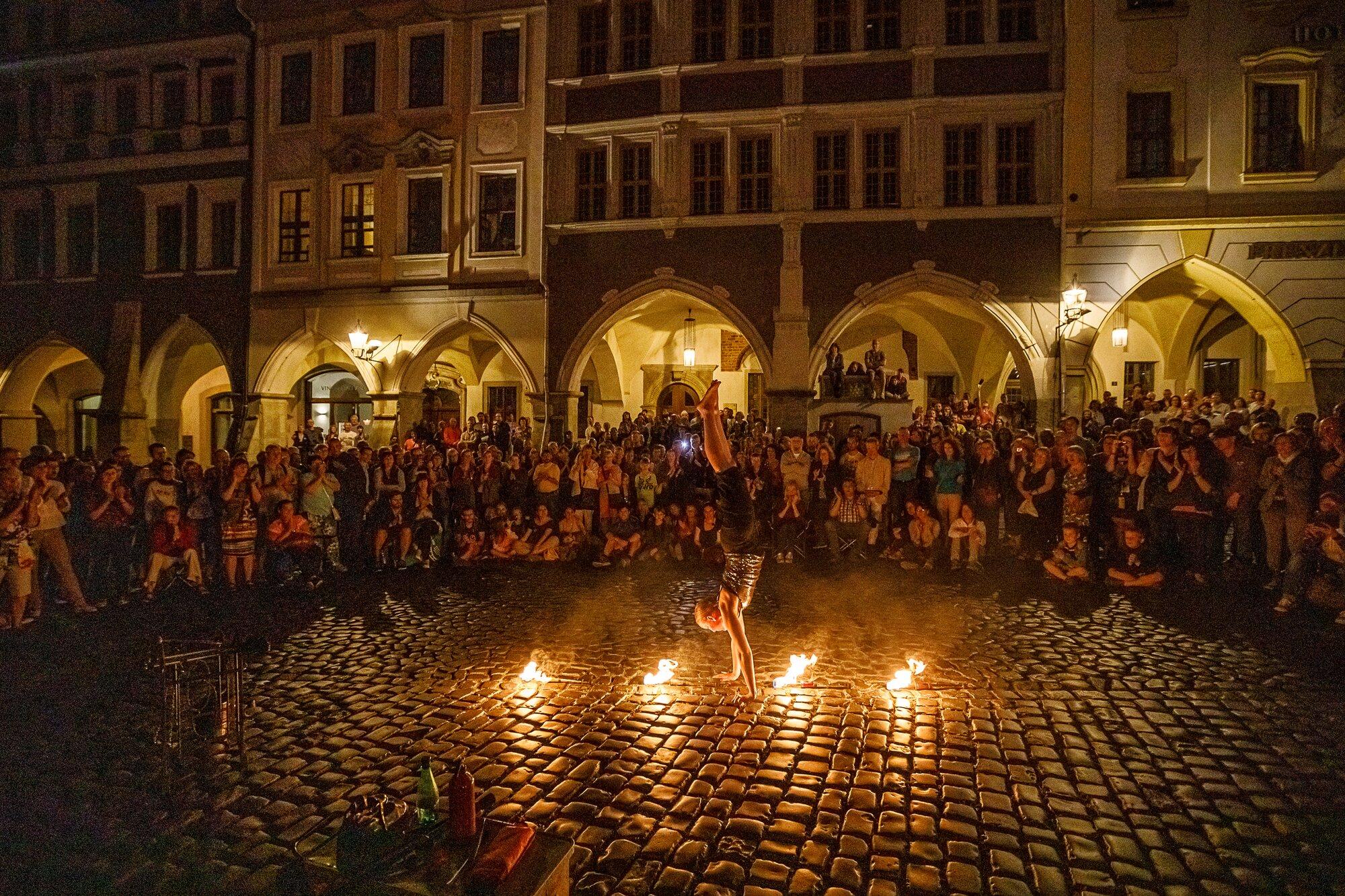 Eine Person macht nachts auf Kopfsteinpflaster einen Handstand zwischen drei brennenden Fackeln. Im Halbkreis dahinter stehen und sitzen viele Zuschauende vor beleuchteten Gebäuden mit Bogengängen.