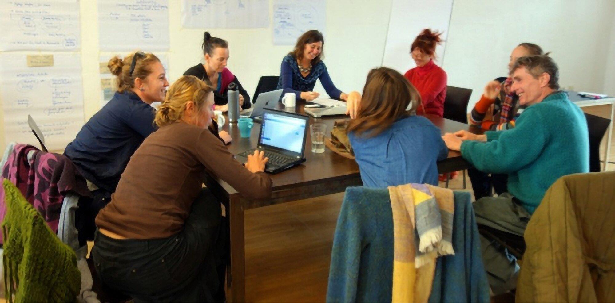 Acht Personen sitzen lachend und im Gespräch um einen großen dunklen Holztisch, darauf Laptops, Tassen und Papiere. An der Wand dahinter hängen beschriftete Flipchart-Bögen, im Vordergrund liegen Jacken über Stuhllehnen.
