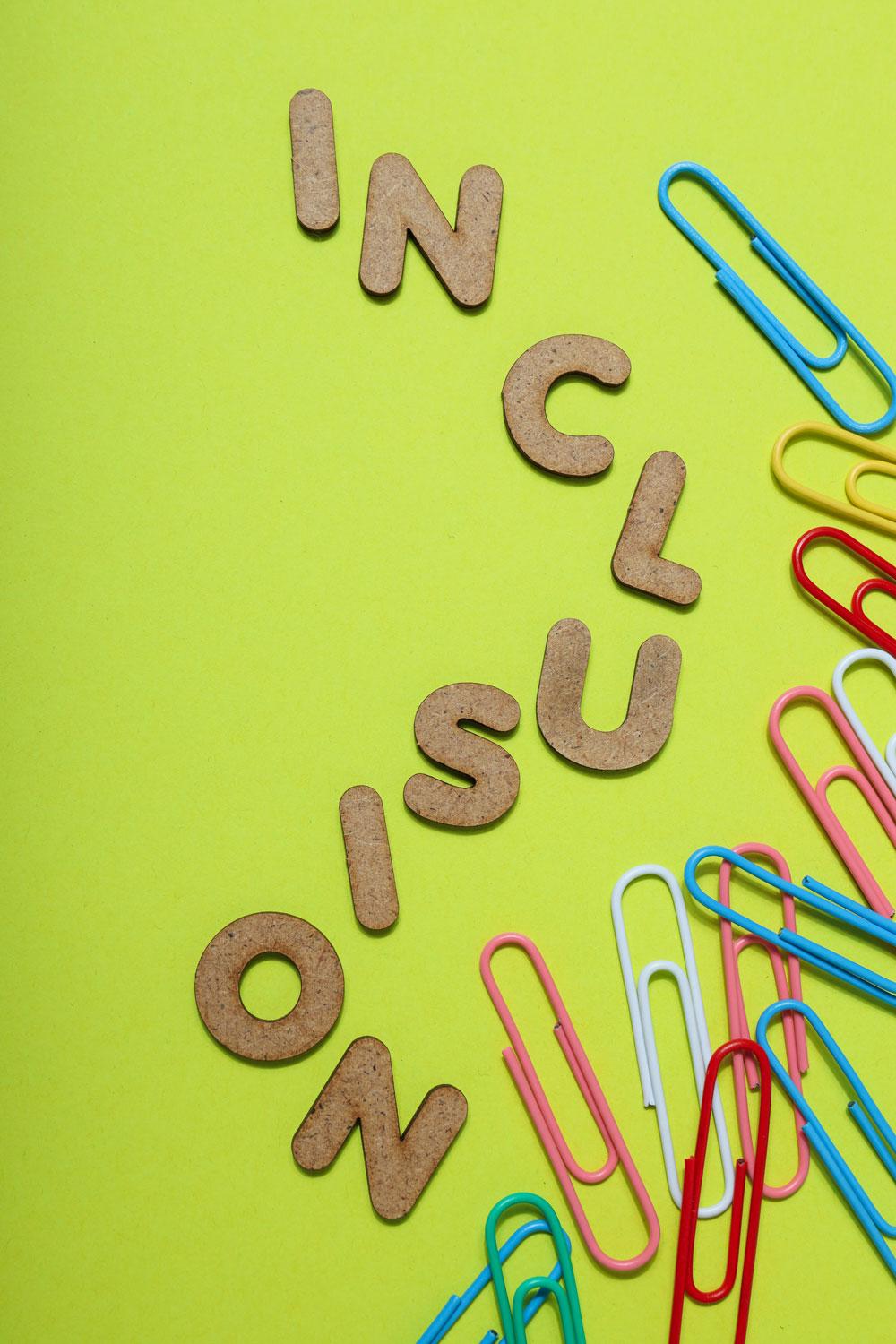 Inklusion-Foto, bestehend aus Buchstaben-Keksen, die "Inclusion" als Wort bilden und bunten Büroklammern auf gelbem Hintergrund.